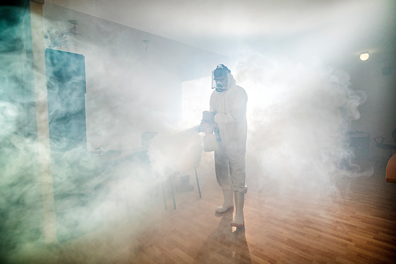 Pest control worker in protective suit fogging a room with thick white smoke indoors