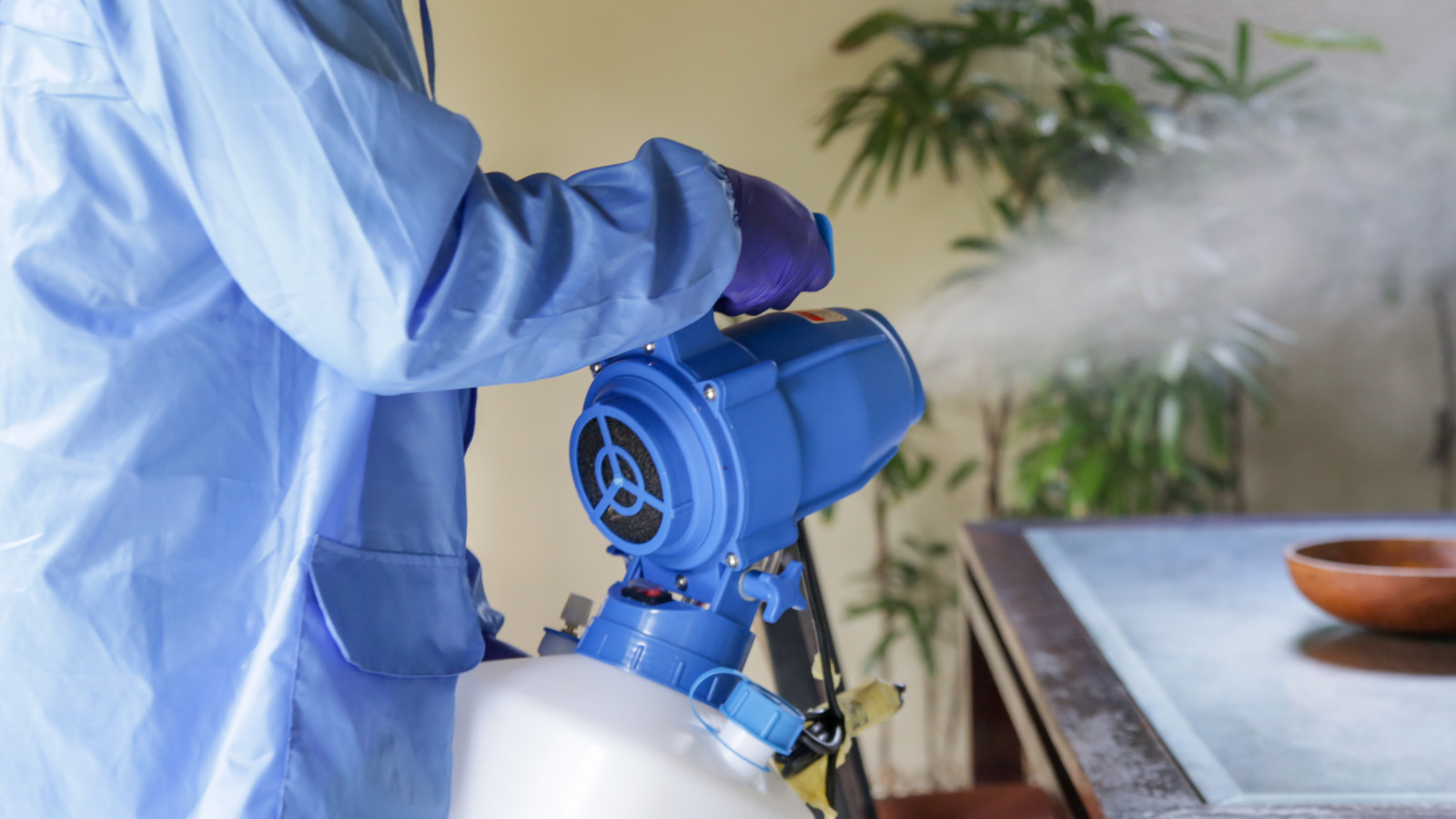 Technician in protective gear using a fumigation spray machine indoors near a table