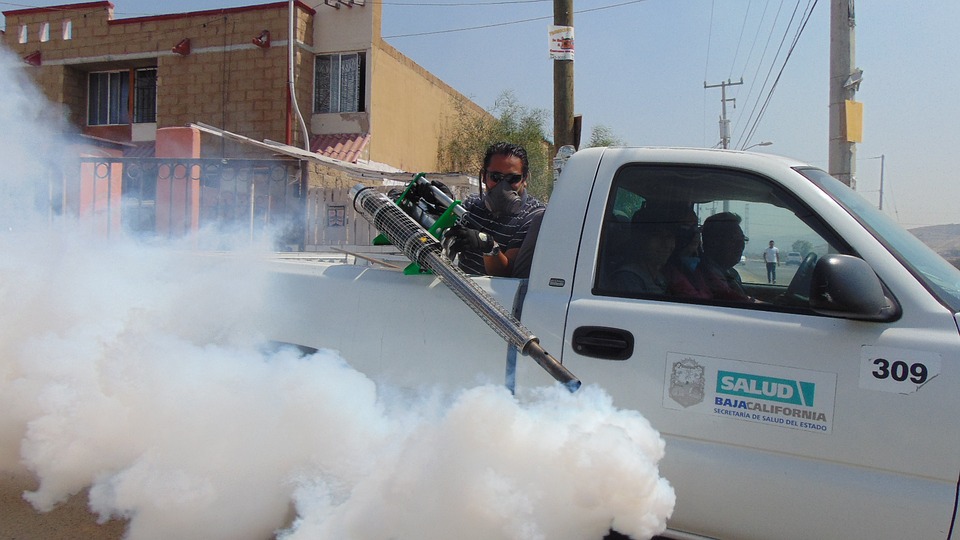 Worker using a fogging machine to spray pesticides from a moving truck in a residential area, producing thick white smoke