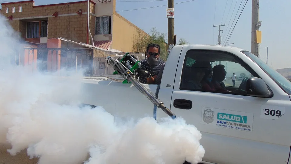 Worker using a fogging machine to spray pesticides from a moving truck in a residential area, producing thick white smoke