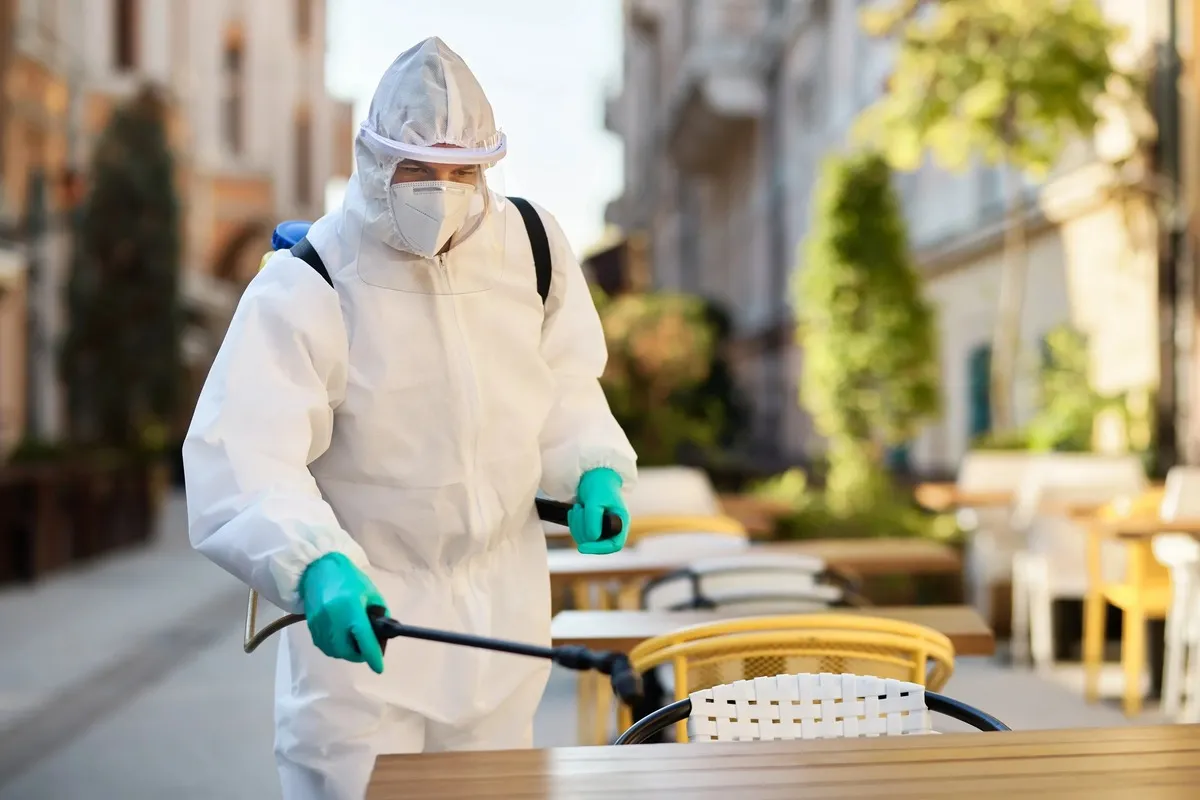 A person wearing a full hazmat suit, face mask, and gloves disinfects outdoor café tables during the COVID-19 pandemic.