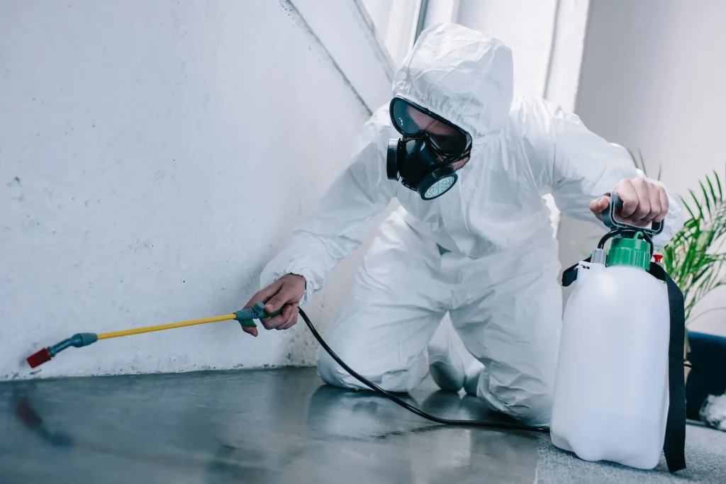 A pest control technician in protective gear applying treatment to a wall to eliminate pests.
