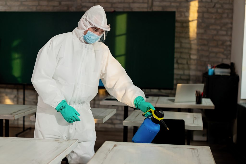 Pest control specialist in hazmat suit spraying school classroom desks with disinfectant – school pest control and sanitization services