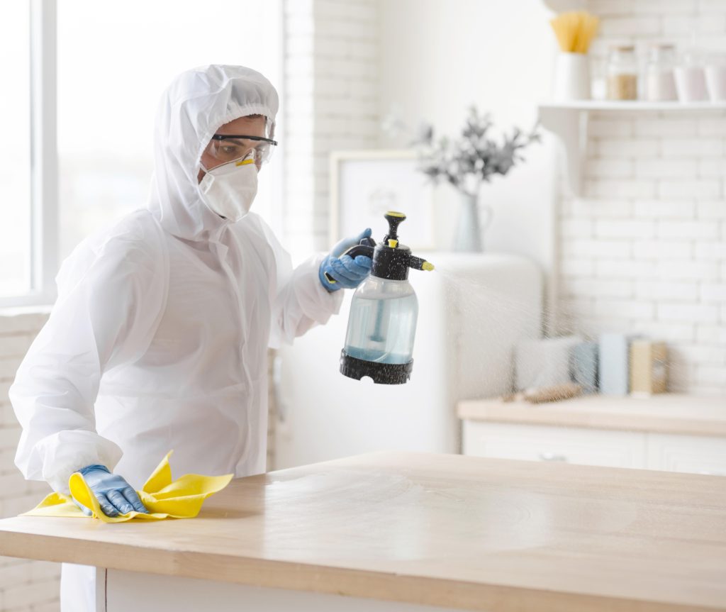 Pest control technician in hazmat suit spraying disinfectant and wiping a wooden table in a hospitality property – hotel pest control treatment