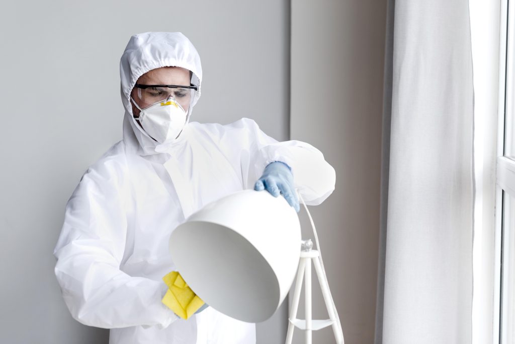 Pest control specialist in protective suit and mask inspecting indoor floor lamp during retail property treatment – retail pest control inspection services