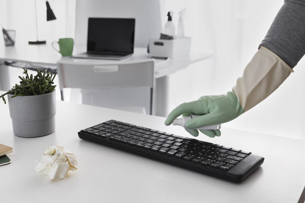 Gloved hand spraying disinfectant on a black keyboard on a white office desk during cleaning process – pest control for offices workstation treatment