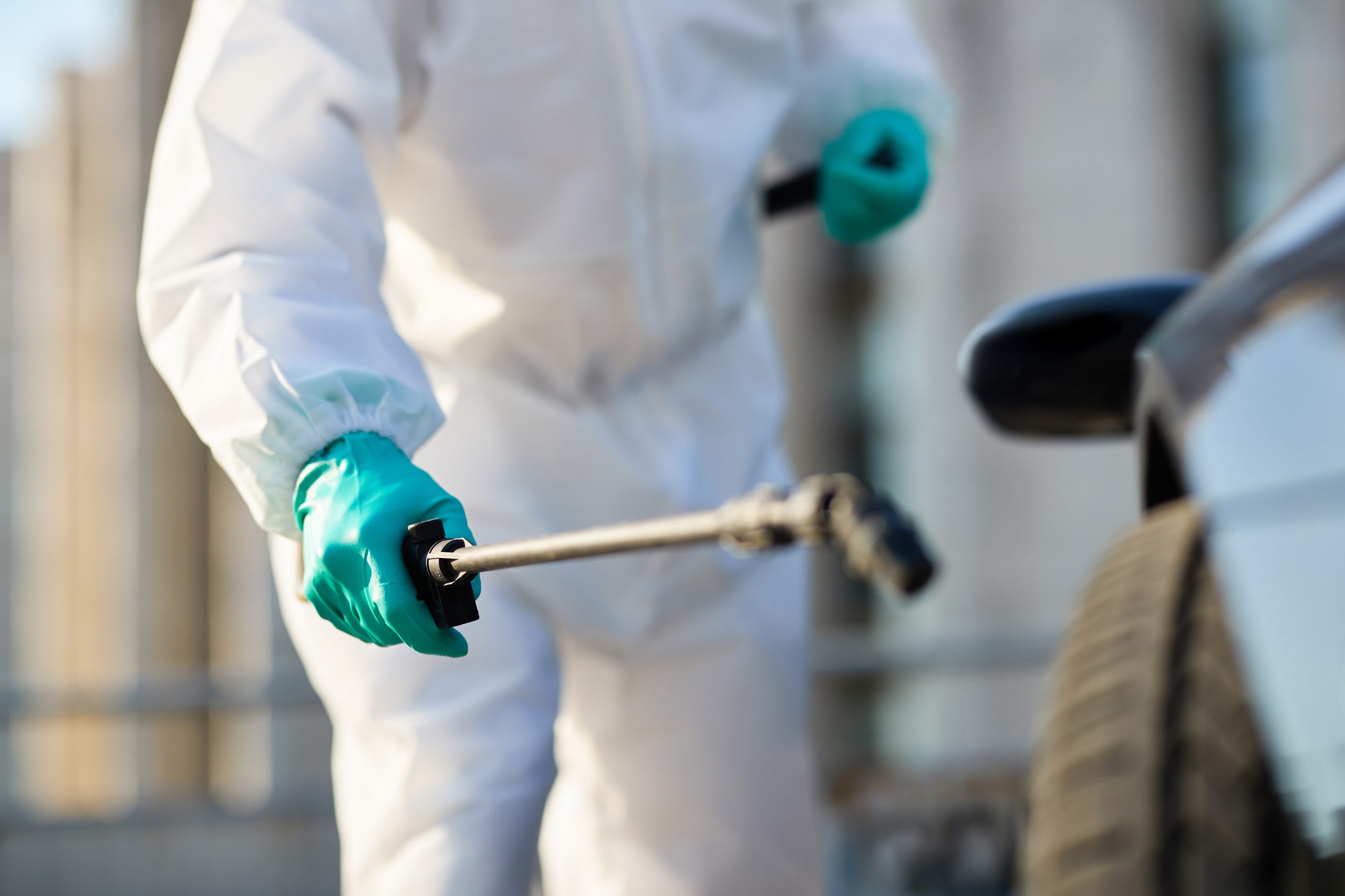 Close-up of pest control technician in white hazmat suit holding spray wand near vehicle – retail pest control treatment services