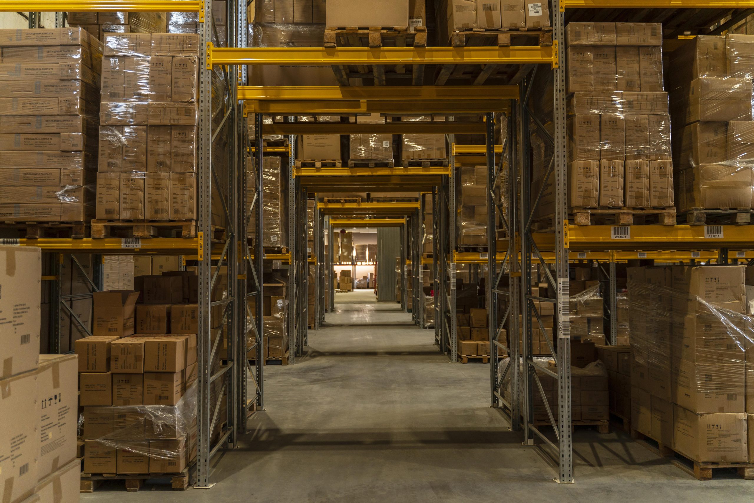 Long aisle of a dark warehouse with tall shelving units stacked with boxed goods on pallets – logistics pest control services