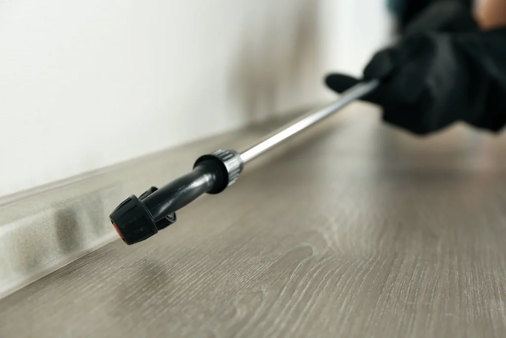 Close-up of pest control spray wand applying treatment along baseboards.
