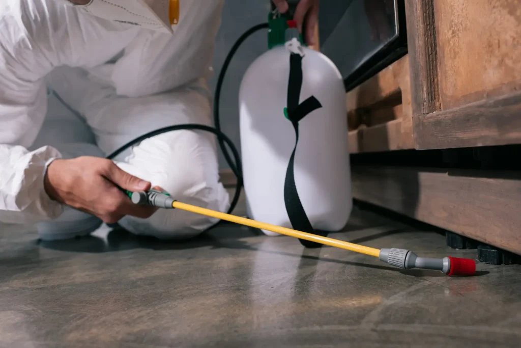 Pest control worker in protective suit spraying insecticide under a cabinet.