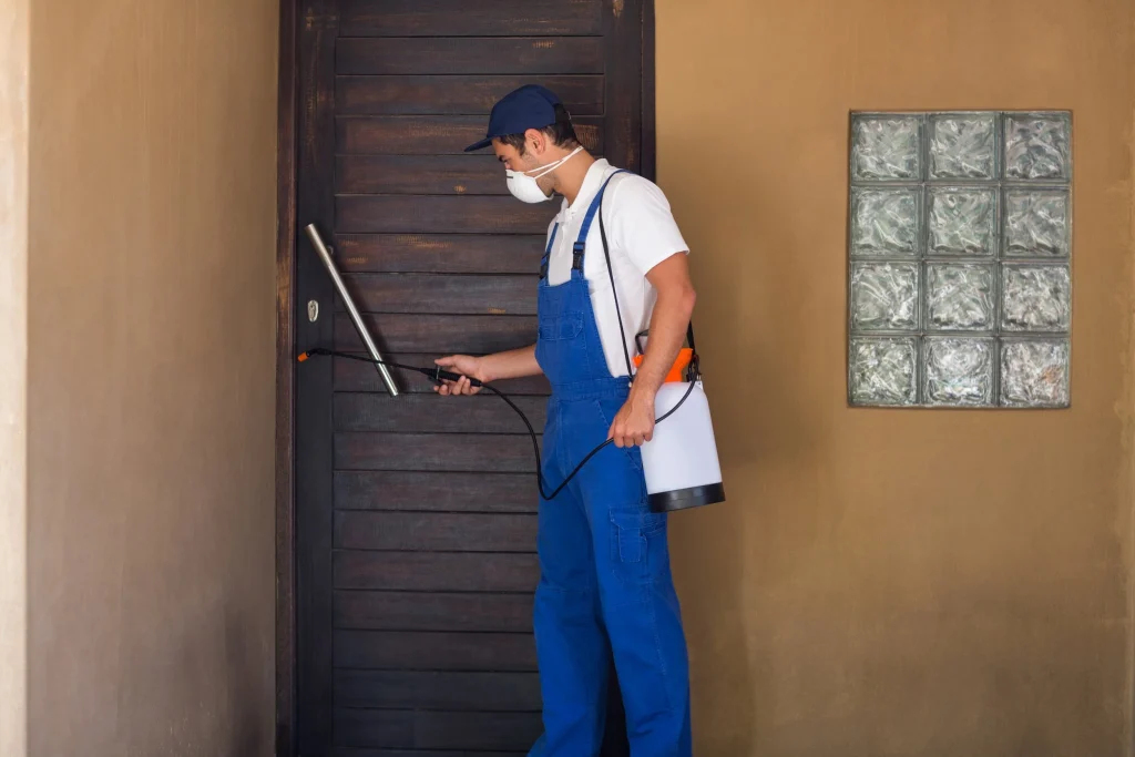 Pest control worker wearing protective gear spraying the exterior of a house.