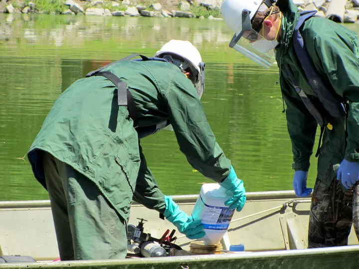 Two people in protective gear are adding chemicals to a boat on a pond for pest safety